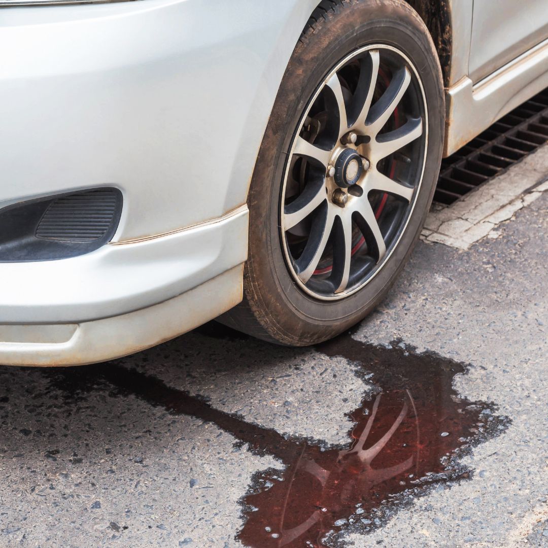 A puddle of reddish-brown fluid is on the ground near the front wheel of a car, indicating a transmission fluid leak.